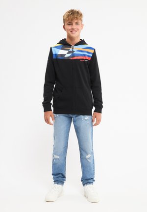 Felpa zip nera con un design geometrico multicolore sulla parte superiore. Caratterizzata da tasche frontali, abbinata a jeans blu chiaro strappati e sneakers bianche.