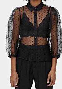 Zwarte doorzichtige blouse met een gestippeld textuur, knoopsluiting aan de voorkant en kraag. Ballonmouwen en een kanten bralette eronder. Geaccentueerd door een getailleerde taille.