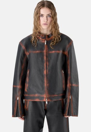 Joven con cabello largo y rizado que lleva una chaqueta y pantalones de cuero negros con detalles desgastados en naranja y cremalleras delanteras.