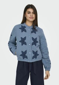 Blå quiltet jakke med marineblå blomsterappliqué, rund krave, frontknaplukning og ribbede manchetter. Båret med mørke bukser.