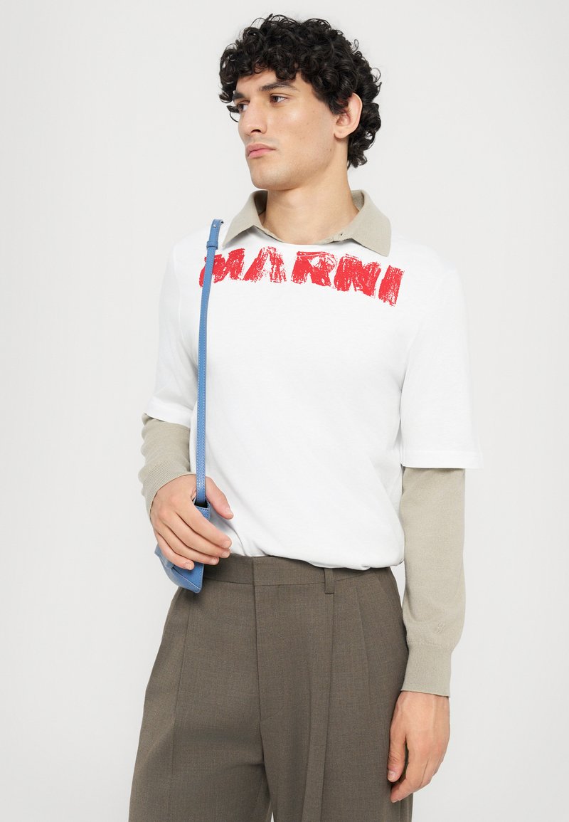 Chemise blanche à manches courtes avec le lettrage rouge "MARNI", col beige, associée à un pantalon gris-brun. Tenant un petit sac à main bleu.