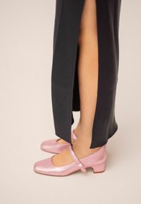 Rosa läder Mary Jane-skor med fyrkantiga tår och låga blockklackar, bärs med en svart klänning med sidofickor, som visar en slät textur.