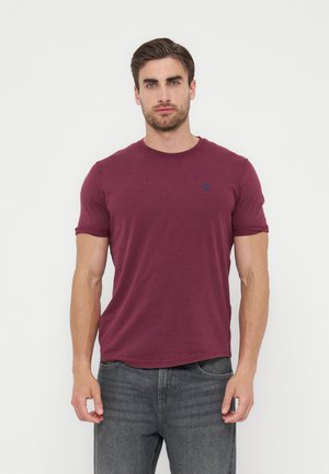 Άνδρας που φοράει μπορντό κοντομάνικο μπλουζάκι και γκρι τζιν, στέκεται μπροστά σε απλό λευκό φόντο, κοιτάζοντας μπροστά με ουδέτερη έκφραση.