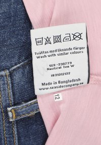 Valkoinen kangas, vaaleanpunainen tausta, jossa hoito-ohjeet. Kiinnitetty tummansinisiin farkkuihin. Sisältää koon etiketin "12" ja bränditiedot.