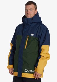 Chaqueta a bloques de color en navy, verde y amarillo. Presenta un alto cuello, bolsillo con cremallera y logo en el pecho. Material impermeable.