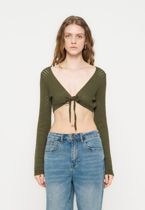 Frau mit langem welligem Haar, die ein olivgrünes gestricktes, kurzes Oberteil trägt, das vorne gebunden ist, und hoch taillierte, ausgewaschene blaue Jeans, stehend vor weißem Hintergrund.