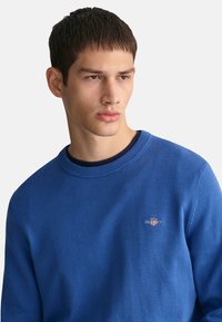 Maglione blu lavorato a maglia con colletto rotondo, dotato di un piccolo logo ricamato sul petto. Il tessuto sembra morbido e ha una texture liscia.