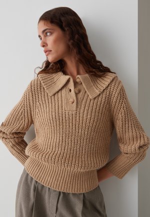 Beige gebreide trui met een grote kraag en een placket met drie knopen. Beschikt over geribbelde manchetten en zoom, weergegeven tegen een lichte achtergrond.