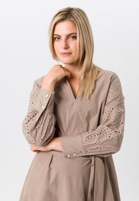 Beige jurk met een V-hals, gedetailleerde ooglets mouwen en knopenaccenten. Heeft een cinched taille met een bijpassende riem.