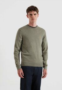 Oliwkowy sweter z okrągłym dekoltem i prążkowanymi mankietami. Posiada małe pomarańczowe logo na piersi. Nosi się go na ciemnych spodniach.