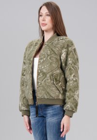 Veste bomber vert olive en fausse fourrure avec une texture douce, fermeture éclair, et poignets côtelés. Elle comprend des poches latérales et une coupe décontractée.
