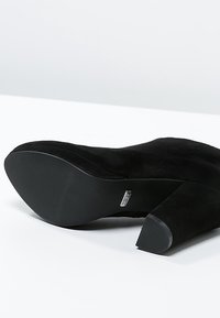 Botín negro de ante con tacón alto, con un tacón grueso, parte superior texturizada y suela suave. Etiqueta del logo visible en el costado.