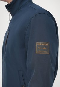 Μπλε σκούρο μπουφάν με ψηλό γιακά. Διαθέτει υφή με ραφές, κλείσιμο με φερμουάρ και καφέ δερμάτινο λογότυπο στο αριστερό μανίκι.