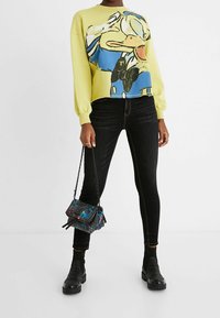 Keltainen neule, jossa sarjakuvakuva, mustat kapeat farkut ja pieni musta käsilaukku monivärisillä kukkakuvioilla. Paksut mustat saappaat.