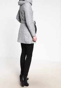 Person klädd i en grå kappa med bälte, svarta leggings och svarta högklackade ankelskor, stående och vänd åt sidan mot en enkel bakgrund.