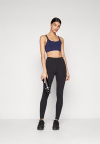 Navy sports bra with thin straps, paired with high-waisted black leggings. Model holds a black water bottle, wearing black athletic shoes.