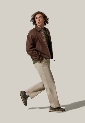 Giacca marrone con cuciture bianche, maglione verde, pantaloni beige e sneakers scure con accenti verdi e marroni. Modello in una posa di camminata.