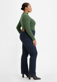 Sweater canelado listrado em verde e azul-marinho, ajustado com gola alta, combinado com jeans de perna larga azul-escuro. Botas de cano curto pretas com salto completam o visual.