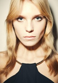 Plavé vlasy s jemnými vlnami rámujú tvár; modré oči, minimálne líčenie; oblečená v čiernom topu so zlatým náhrdelníkom.