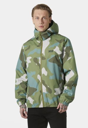 Mężczyzna z tatuażami ubrany w zieloną, niebieską i białą kamuflażową kurtkę z kapturem na suwak oraz czarne spodnie, stojący na białym tle.