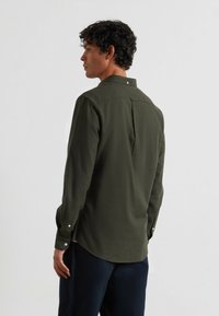 Ciemnozielona koszula z długim rękawem, wykonana z teksturowanej tkaniny. Posiada kołnierzyk zapinany na guziki, widoczne szwy oraz rozcięcia po bokach u dołu.