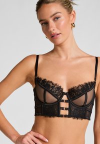 Μαύρο σουτιέν με διάφανο διχτυωτό σχέδιο και λεπτομέρειες από δαν�τέλα. Οι θήκες με ενίσχυση έχουν φινίρισμα με κυματιστό άκρο και διακοσμητικά αξεσουάρ μπροστά.