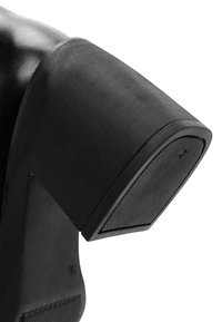 Scarpa nera con tacco alto, caratterizzata da un tacco largo e texture. Il materiale sembra lucido, con una suola in gomma dotata di una presa a motivo.