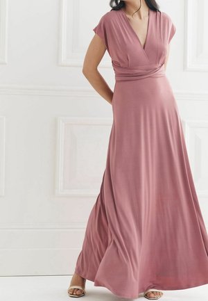 Kvinde iført en støvet rosafarvet, gulvlængde slå-om-kjole med korte ærmer og beige, åbne højhælede sko foran en hvid væg med paneler.