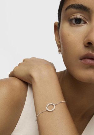 Kobieta opierająca rękę na ramieniu, nosząca delikatną srebrną bransoletkę z okrągłym zawieszkiem oraz dopasowaną małą kołową kolczyk.