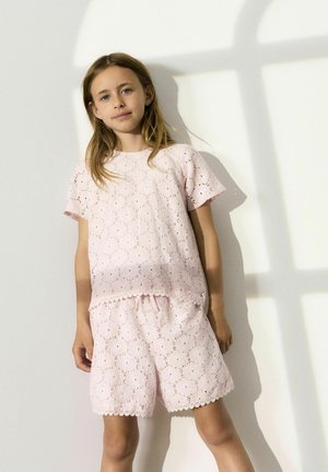 Completo in pizzo di colore rosa chiaro, composto da una maglietta a maniche corte e pantaloni corti con vita elastica. Motivo floreale intricato con bordi scallop.