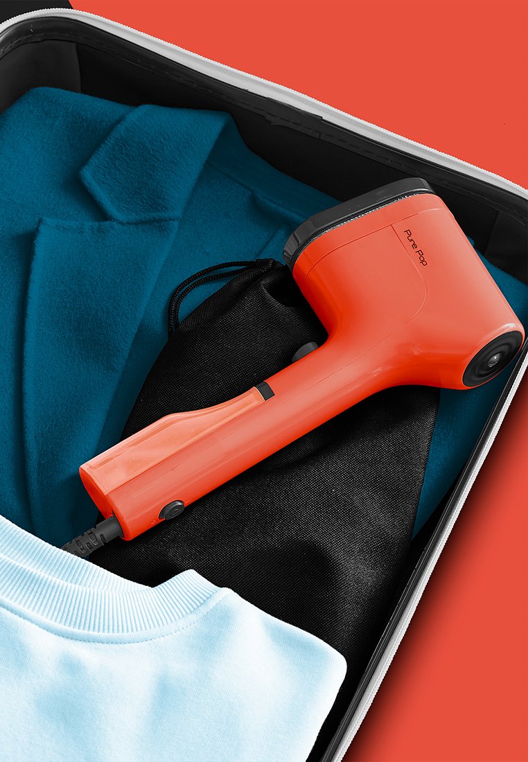 Compact oranje kledingstomer met een zwart waterreservoir, rustend op blauwe stof, naast een lichtblauwe kraag en een zwarte tas in een koffer.