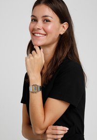 Montre numérique avec un cadran rectangulaire doré, un bracelet en métal argenté et des accents noirs. L'affichage montre l'heure au format numérique.
