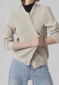 Beige bomberjack met een ritssluiting aan de voorkant, ribgebreide manchetten en zoom. Gladde textuur, aansluitend ontwerp, gedragen over een zwart topje en blauwe jeans.