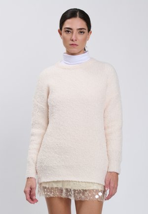 Woman with dark hair pulled back wearing a white fuzzy sweater over a sheer beige skirt with star patterns against a plain light background.
