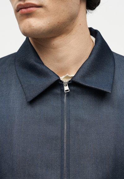 Κοντινό πλάνο ενός ατόμου που φοράει σκούρο μπλε ζακέτα με φερμουάρ και γιακά πάνω από ένα ανοιχτόχρωμο πουκάμισο, φαίνεται από τον λαιμό μέχρι το μέσο του στήθους.