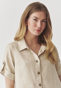 Camicia in lino beige con colletto, maniche arrotolate e bottoni in legno. Texture liscia con una finitura naturale.