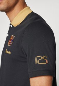 Polo nero con colletto dorato, caratterizzato da logo ricamato del Barcellona, testo "Spotify" e un design dorato "PS" sulla manica.