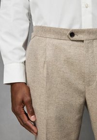 Pantalon beige à chevrons avec une coupe ajustée, doté de poches latérales, d'une fermeture à bouton noir unique et d'une texture de tissu lisse.
