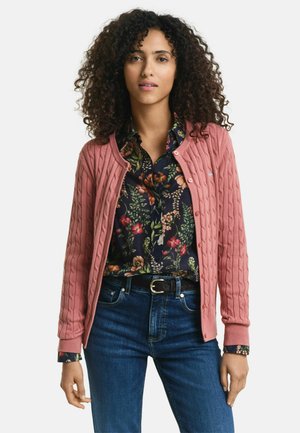 Roze kabelgebreide cardigan met een ronde halslijn en knoopsluiting, gelaagd over een bloempjeshemd, gedragen met donkere jeans.