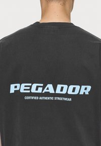 Černé tričko s velkým světle modrým potiskem na zádech, na kterém je napsáno "PEGADOR" a pod tím "CERTIFIED AUTHENTIC STREETWEAR".