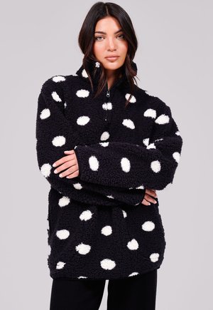 Femme portant un pull polaire noir avec des pois blancs, debout les bras croisés devant un fond clair uni.