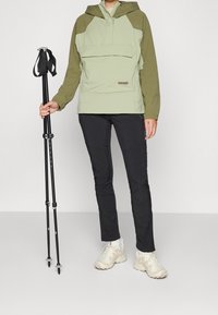 Sudadera verde y oliva con capucha, bolsillo frontal y pantalones negros. Bastones de trekking negros y zapatillas beige completan el conjunto.