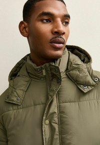 Chaqueta acolchada de color verde oliva con cuello alto, cremallera y botones a presión. Presenta una superficie texturizada y una capucha con detalles de botones a presión.