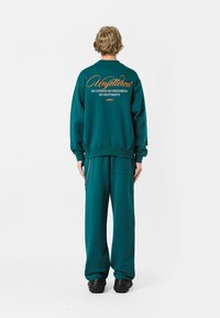 Sudadera color verde azulado con el texto "Unfettered" y eslogan en la parte posterior, combinada con pantalones a juego. Fabricada en material suave y con un ajuste relajado.