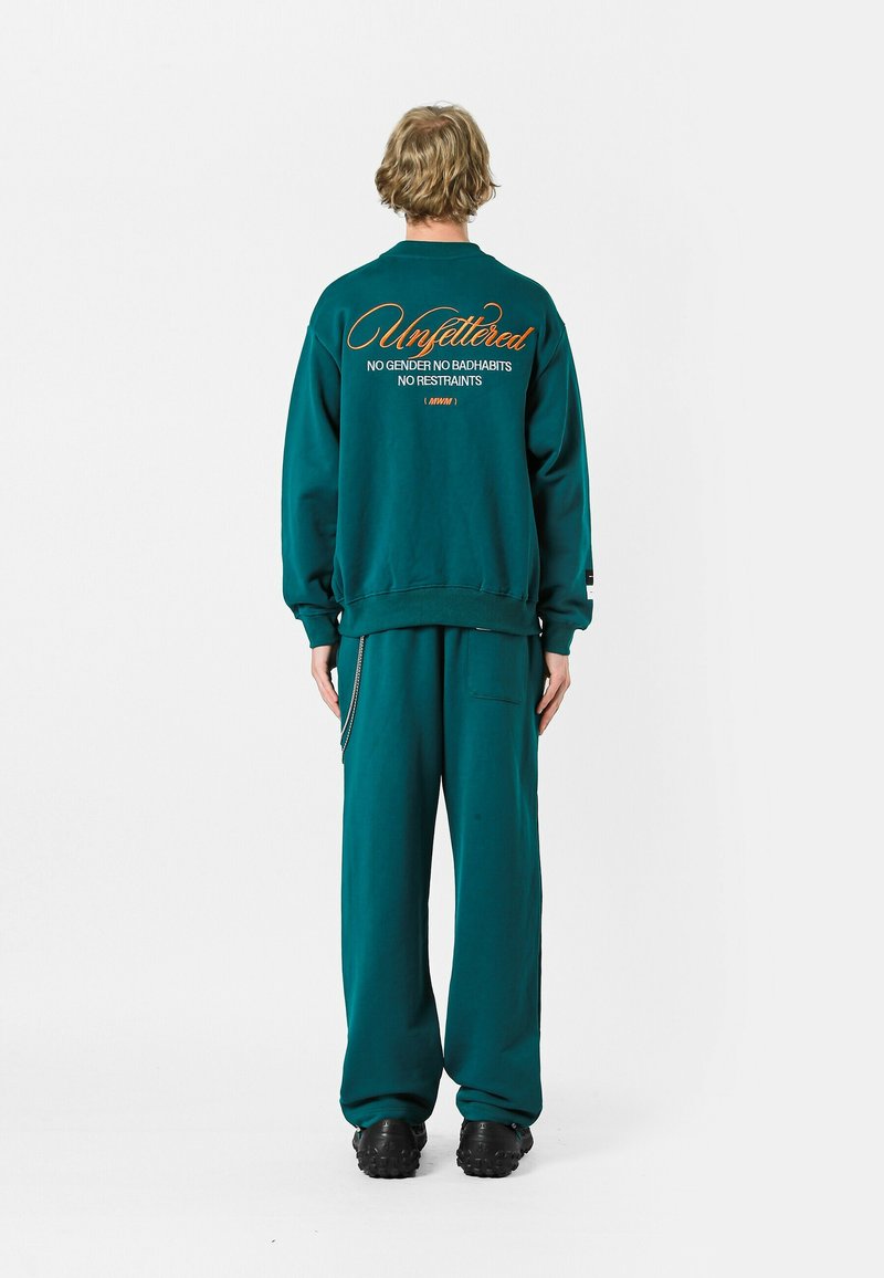 Sudadera color verde azulado con el texto "Unfettered" y eslogan en la parte posterior, combinada con pantalones a juego. Fabricada en material suave y con un ajuste relajado.