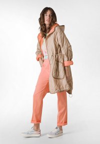 Chaqueta acolchada beige con capucha naranja, camiseta gráfica rosa claro y pantalones coral. Zapatillas blancas con acentos en azul y rosa completan el conjunto.
