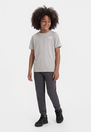 Χαμογελαστό παιδί με σγουρά μαλλιά, που φοράει ανοιχτό γκρι κοντομάνικο μπλουζάκι, σκούρο γκρι παντελόνι και μαύρα αθλητικά παπούτσια, στέκεται μπροστά σε λευκό φόντο.