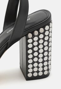 Crne sandale s visokom potpeticom i debelim blok petom ukrašene srebrnim zakovicama. Imaju elegantne kožne trake i strukturiranu ulošak.