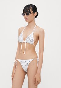 Biely bikiny set s trojuholníkovými pohármi a kovovými ozdobami. Tká na krku a bokoch, s hladkou textúrou a minimalistickým dizajnom.