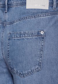 Vaqueros de mezclilla azul que presentan un bolsillo trasero con un detalle de botón, textura desgastada y una etiqueta rectangular con texto en relieve.